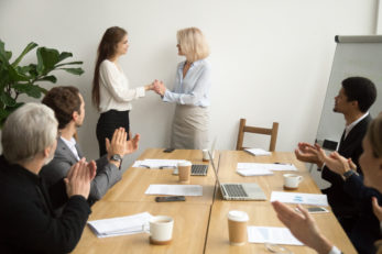 Senior businesswoman boss promoting female employee thanking for good work holding hands as trust and support concept while team applauding congratulating colleague at group meeting, recognition